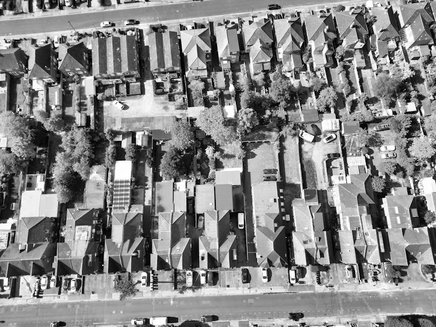 An aerial black-and-white photograph of a residential neighbourhood with rows of closely packed terraced houses, each with pitched roofs and small front gardens. The houses are separated by narrow alleyways and backyards, some containing trees, utility sheds, or exercise equipment. Several parked cars are visible along the streets and in driveways. In the foreground, a wider street runs horizontally, with vehicles parked along its edges and sidewalks on either side. The image depicts a typical urban area suitable for home relocation projects, where removals services such as furniture transport, packing, and loading are often required. The detailed view of the houses and surrounding environment highlights the importance of careful planning in moving logistics, including navigating access issues on Kennington Park Road, as referenced in the page title, with [COMPANY_NAME] providing professional removals solutions.