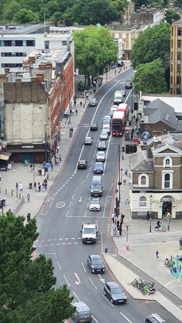 An aerial view of Kennington Park Road showing a narrow urban street lined with various buildings, including residential houses with pitched roofs and commercial premises. The road has multiple lanes with cars and vans stationary in a queue, suggesting a temporary hold during a home relocation or furniture transport process. Sidewalks on both sides are populated with pedestrians, some walking and others waiting, and bicycles are parked near the sidewalk. Tall, leafy green trees shade parts of the street, and a large truck or van is positioned close to the curb, likely involved in an ongoing moving or packing operation. The scene is illuminated by daylight, capturing the typical hustle of street-level vehicle loading and pedestrian activity associated with relocation services in an out-of-town residential area. This visual reflects aspects of moving logistics, transport, and packing processes that are relevant to removals specialists like Removals Kennington.
