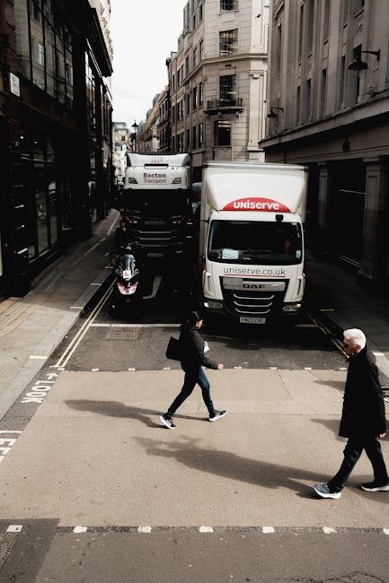 A street scene showing a vehicle loading area with two white moving vans parked parallel to the surroundings, one branded with 'Uniserve' on the front and sides, and a smaller van with 'Becton Transport' on its rear. The vans are positioned close to a narrow alley or loading zone between tall, multi-storey buildings featuring classic architectural details such as fire escapes and large windows. In the foreground, a man with grey hair dressed in a dark coat and trousers is walking on the pavement, while a woman wearing casual clothes and holding a bag crosses the street nearby. The area appears to be in a busy urban setting, with the loading process indicating a house removal or home relocation activity supported by [COMPANY_NAME], with vehicles and personnel involved in furniture transport and packing procedures. The scene is captured in natural daylight, providing clear visibility of the vehicles, the loading environment, and the pedestrians involved in the moving operation.