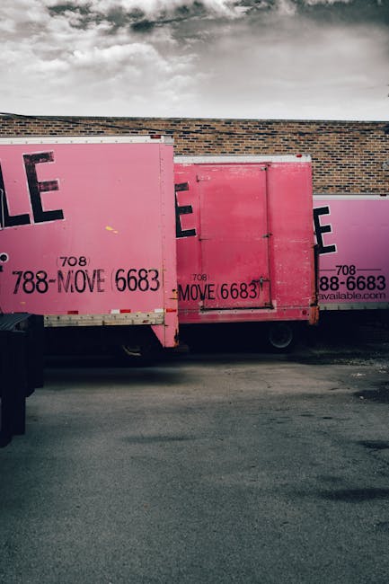 The image shows three large pink moving containers positioned on a paved surface adjacent to a brick building with a hill and cloudy sky in the background. The containers are made of metal with pink painted exteriors, and they feature large black text displaying contact details and service information for house removals, including phone numbers and website details. The containers are part of a home relocation process, likely used for packing or transporting furniture, appliances, and household items. One container has a visible side door, and the containers are closely situated, suggesting they are either being loaded onto a vehicle or prepared for moving activities. There are no people visible in the image, but the scene indicates an active loading or unloading operation consistent with furniture transport or packing during a house removal project. Removals Kennington offers such services, and these containers are typical of the bulk packaging used in professional moves.