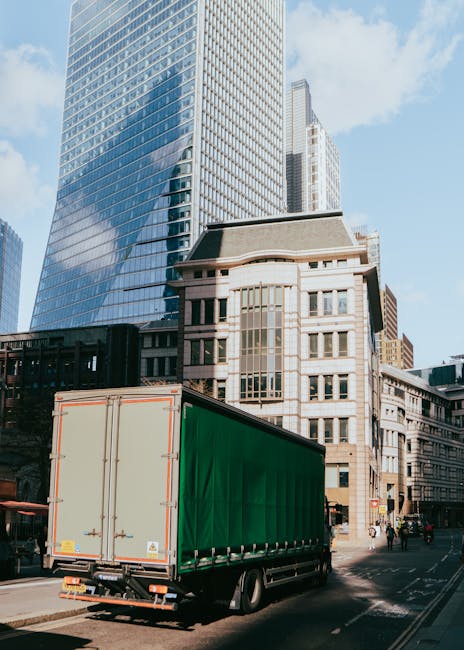 A street scene showing a vehicle loading area with two white moving vans parked parallel to the surroundings, one branded with 'Uniserve' on the front and sides, and a smaller van with 'Becton Transport' on its rear. The vans are positioned close to a narrow alley or loading zone between tall, multi-storey buildings featuring classic architectural details such as fire escapes and large windows. In the foreground, a man with grey hair dressed in a dark coat and trousers is walking on the pavement, while a woman wearing casual clothes and holding a bag crosses the street nearby. The area appears to be in a busy urban setting, with the loading process indicating a house removal or home relocation activity supported by [COMPANY_NAME], with vehicles and personnel involved in furniture transport and packing procedures. The scene is captured in natural daylight, providing clear visibility of the vehicles, the loading environment, and the pedestrians involved in the moving operation.
