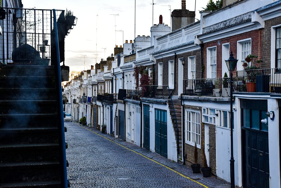 A narrow residential street lined with terraced houses featuring brick and white facades, with small front gardens and balconies equipped with black metal railings. The street is paved with cobblestones, and some houses have potted plants and outdoor lighting fixtures on their exteriors. On the left side, a black metal staircase with steps made of the same material ascends from the pavement to a higher level, partially visible against the sky, which appears overcast with soft natural light. The scene illustrates a typical urban setting suitable for home relocation or furniture transport services, with clear access points for moving companies like Removals Kennington. The image captures an environment where loading and unloading personal belongings or packing materials could take place during a house removal process, highlighting the importance of careful planning for moving logistics in such a setting.