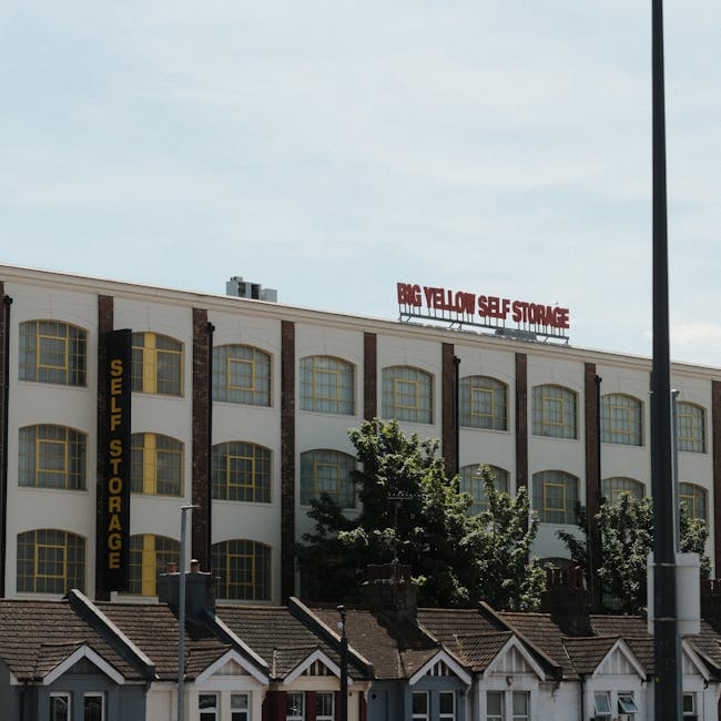 Image of a multi-storey commercial building with a large sign reading 'BIG YELLOW SELF STORAGE' in red letters on the roof, and a prominent vertical black sign on the left side that says 'SELF STORAGE' in yellow letters. The building features multiple large, rectangular yellow-framed windows, some with yellow curtains, and brick columns between the window sections. In the foreground, there are small residential-style roofed structures with white frames, likely part of a residential neighbourhood, and a street lamp at the right edge of the image. The scene is taken during daylight with a partly cloudy sky, illustrating the location used for home relocation and furniture transport storage by removals Kennington, supporting the process of packing and moving.