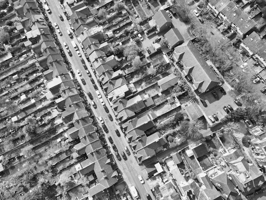 An aerial black-and-white photograph of a residential street showing closely packed terraced houses with pitched roofs, front and back gardens, and lined-up parked cars along both sides of the narrow road. The street features a row of houses on each side, separated by a central road with vehicles parked in designated spaces. Trees and shrubs are visible between some properties, and the scene includes a few pedestrians walking on the pavement. The image captures the typical urban layout of a neighbourhood with dense housing, with some houses having small driveways or garden areas. The photograph appears to be taken during daylight with natural lighting and shadows visible on rooftops and streets. This image relates to house removals and furniture transport, illustrating the environment where [COMPANY_NAME] conducts home relocation services, including packing, loading, and transportation of household belongings involved in moving processes.