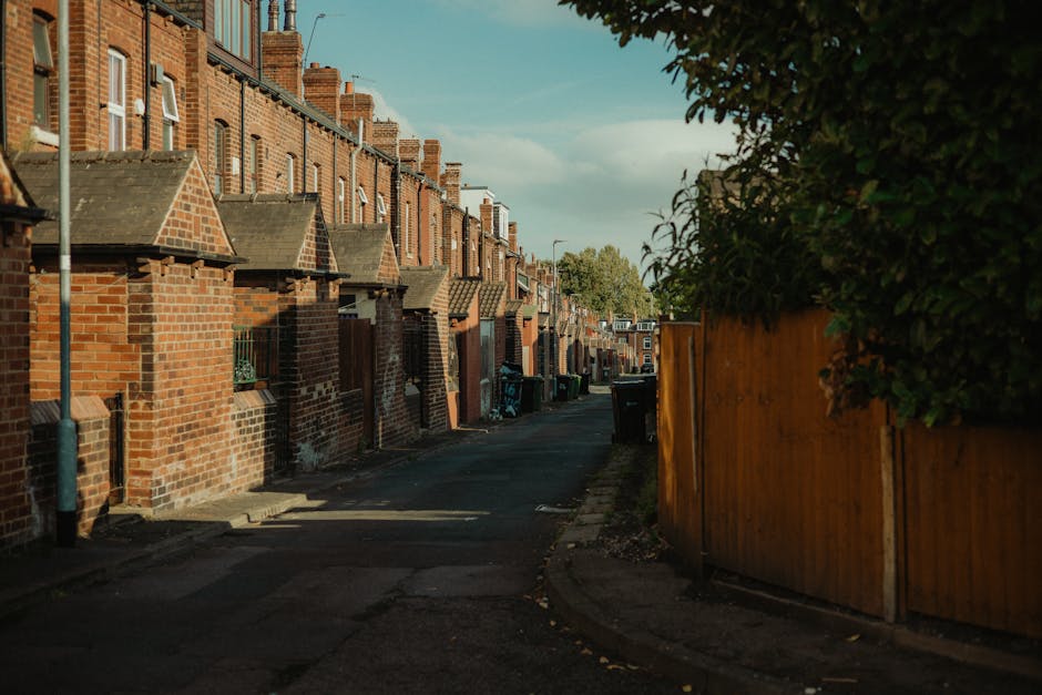 A quiet residential backstreet in Kennington with a row of terraced brick houses on the left, featuring small front steps and pitched roofs. The street is narrow, paved with asphalt, and lined with various black and green wheelie bins positioned next to the houses, indicating collection points. On the right, a tall wooden fence runs parallel to the street, partially obscuring a garden area with dense green foliage. The scene is lit with soft natural light, suggesting late afternoon, and there are no vehicles or pedestrians visible. This setting exemplifies the typical environment where Removals Kennington might facilitate home relocation or furniture transport, with a focus on loading and moving processes within such urban residential areas.
