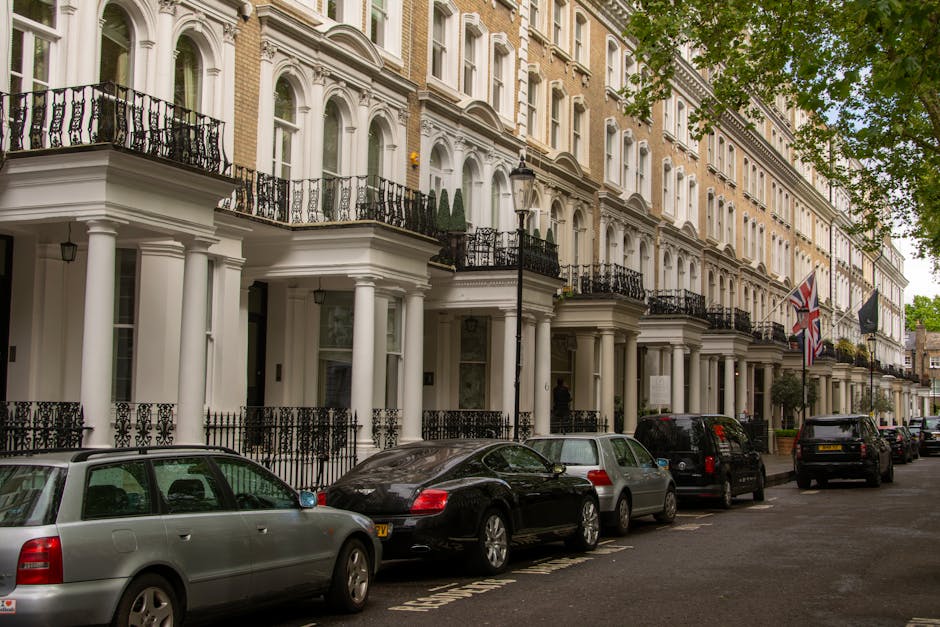 An aerial black-and-white photograph of a residential street showing closely packed terraced houses with pitched roofs, front and back gardens, and lined-up parked cars along both sides of the narrow road. The street features a row of houses on each side, separated by a central road with vehicles parked in designated spaces. Trees and shrubs are visible between some properties, and the scene includes a few pedestrians walking on the pavement. The image captures the typical urban layout of a neighbourhood with dense housing, with some houses having small driveways or garden areas. The photograph appears to be taken during daylight with natural lighting and shadows visible on rooftops and streets. This image relates to house removals and furniture transport, illustrating the environment where [COMPANY_NAME] conducts home relocation services, including packing, loading, and transportation of household belongings involved in moving processes.