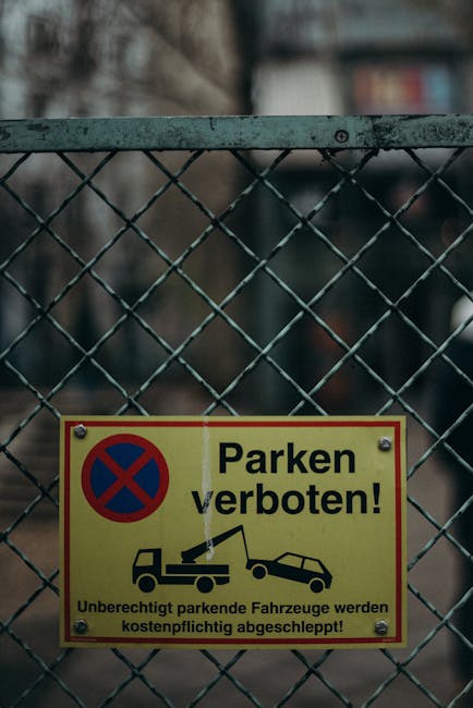 A close-up of a yellow and red rectangular sign attached to a metal diamond-patterned fence, displaying a blue circle with a red border and an 'X' symbol indicating no parking. The sign features black text in German that reads 'Parken verboten!' and includes black pictograms illustrating a car being towed by a crane, with additional smaller text warning that illegally parked vehicles will be removed at the owner's expense. The fence is made of metal with a weathered appearance and is situated outdoors, with blurred background elements suggesting an urban environment. This image contextualizes strict parking restrictions, relevant to house removals and transportation services by Removals Kennington, highlighting the importance of adhering to local parking rules during home relocation and furniture transport processes.