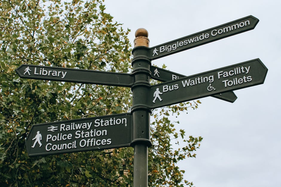 A black multi-directional street sign post situated outdoors against a backdrop of green-leafed trees and an overcast sky. The signpost features several rectangular signs with white text and icons, providing directions to local amenities such as Biggleswade Common, the library, railway station, police station, council offices, bus waiting facility, and toilets, including accessible facilities. The signs are mounted on a central pole with metal brackets, with some signs pointed horizontally while others are angled slightly upward. The wayfinding signage is clear and legible, serving pedestrians and visitors navigating the area. This image relates to local navigation and is relevant to house removals or home relocation services in the Kennington area, highlighting the importance of clear signage during the moving process, especially when planning transport or packing activities involved in a home move with companies like Removals Kennington.