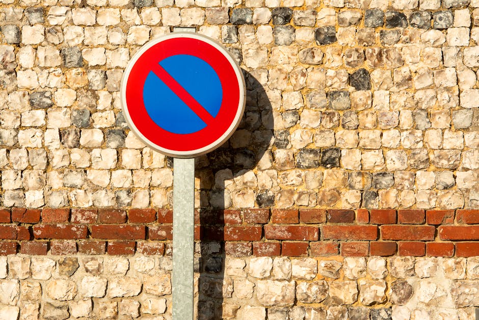 A close-up of a yellow and red rectangular sign attached to a metal diamond-patterned fence, displaying a blue circle with a red border and an 'X' symbol indicating no parking. The sign features black text in German that reads 'Parken verboten!' and includes black pictograms illustrating a car being towed by a crane, with additional smaller text warning that illegally parked vehicles will be removed at the owner's expense. The fence is made of metal with a weathered appearance and is situated outdoors, with blurred background elements suggesting an urban environment. This image contextualizes strict parking restrictions, relevant to house removals and transportation services by Removals Kennington, highlighting the importance of adhering to local parking rules during home relocation and furniture transport processes.