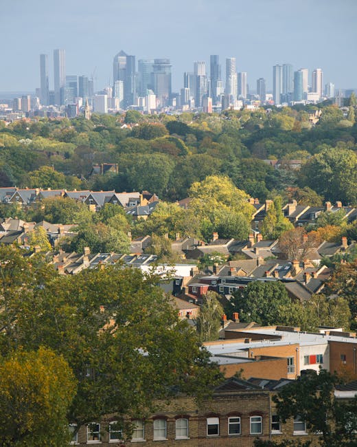 A panoramic view of a residential neighbourhood with tree-lined streets and rooftops in the foreground, leading to a distant city skyline featuring modern high-rise office buildings and skyscrapers. The scene is viewed from an elevated position, possibly from a hill or upper floor window, during daylight with natural lighting. The image showcases a contrast between the suburban houses with pitched roofs and the urban towers in the background, highlighting the space for house removals and moving services near Kennington. The presence of greenery and the detailed rooftops provide context for relocation activities such as packing, loading, and transportation planning by companies like Removals Kennington, which specialises in furniture transport and home relocation services within the London area. The overall scene captures the transition from residential to commercial environments, emphasizing the logistical aspects involved in house removals, including transport routes and urban planning considerations.