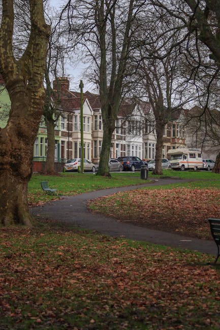 An outdoor scene in a residential area during daytime, featuring a paved walkway winding through a park with fallen autumn leaves scattered on the ground. Two empty metal benches are positioned on either side of the path, and several large, leafless trees with thick trunks and sprawling branches are visible, casting shadows across the park. In the background, a row of Victorian-style terraced houses with bay windows and decorative brickwork is seen along a street, with parked cars including a white van, a silver hatchback, and a black estate car. The scene appears to depict the entrance or vicinity of a house removal or relocation process, with the park providing a tranquil setting close to the homes, relevant to [PAGE_TITLE] and serviced by [COMPANY_NAME]. The lighting is natural and subdued, consistent with an overcast day, enhancing the clarity of the elements within the scene, including the parked vehicles, trees, and park features.