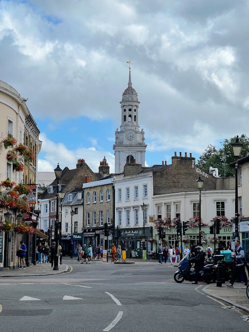 A busy street scene in a historic town with multi-story buildings featuring classic architectural details, including large windows and decorative facades. In the background, a tall white clock tower with a spire and a weather vane stands prominently against a partly cloudy sky. The street is populated with pedestrians, some walking and others standing outside shops, and a mix of vehicles including motorcycles and cars. Alongside the buildings, there are street lamps and hanging flower baskets adding colour and charm. The scene captures the bustling atmosphere typical of a home relocation area, with people moving around near shop entrances and a sense of organized activity suggesting a relocation service like Removals Kennington handling furniture transport and packing in the vicinity, supporting the visual narrative of a busy urban area suitable for same-day removals near Kennington Oval.