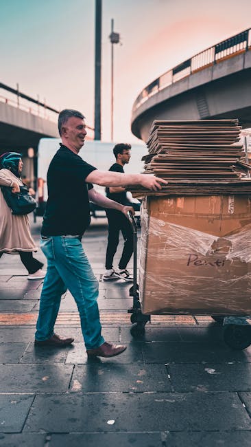 A man wearing a black shirt and blue jeans is pushing a large wheeled trolley loaded with multiple stacked cardboard boxes and packing materials, some wrapped in plastic film, on a city pavement. Behind him, several other people are engaged in what appears to be a home relocation or moving process near a modern bridge structure and streetlights. The scene is set during dusk or evening, with soft lighting highlighting the urban environment. The trolley and stacked boxes indicate the process of furniture transport and packing as part of a professional removals operation, possibly coordinated by Removals Kennington. The image captures the logistical activity involved in loading and transporting belongings for house removals, emphasizing careful handling and organized moving practices in an outdoor setting.
