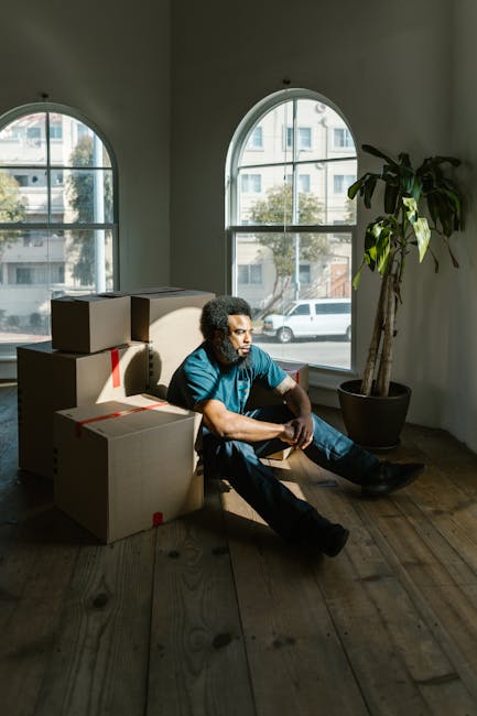 A man with curly dark hair and wearing a teal shirt and dark trousers is sitting on a wooden floor inside a room with two large arched windows, through which a residential street with parked cars and trees is visible. The man appears to be resting amidst several cardboard boxes that are stacked around him, some sealed with red tape, indicating they are packed for a home relocation. The room features a potted plant next to the window, adding a touch of greenery, and the sunlight streaming through the windows casts shadows on the walls and floor. This scene captures the preparation stage of packing and moving, with the boxes ready for transport, and the man taking a moment during the loading process, which is part of the services offered by Removals Kennington. The image reflects the key elements involved in furniture transport and packing during house removals.