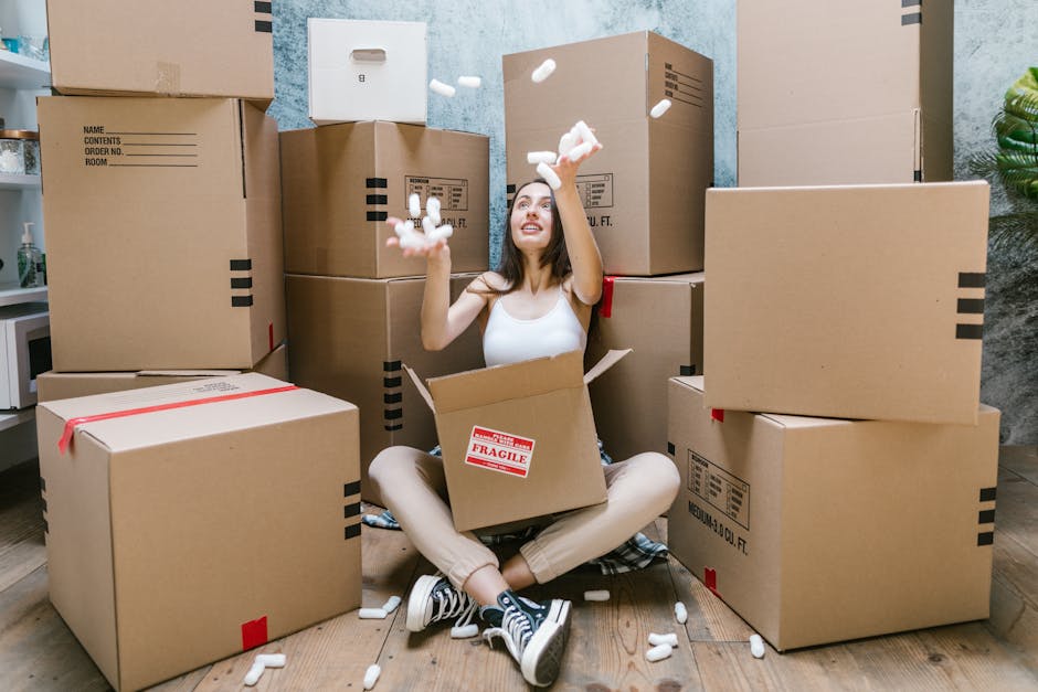 A man with curly dark hair and wearing a teal shirt and dark trousers is sitting on a wooden floor inside a room with two large arched windows, through which a residential street with parked cars and trees is visible. The man appears to be resting amidst several cardboard boxes that are stacked around him, some sealed with red tape, indicating they are packed for a home relocation. The room features a potted plant next to the window, adding a touch of greenery, and the sunlight streaming through the windows casts shadows on the walls and floor. This scene captures the preparation stage of packing and moving, with the boxes ready for transport, and the man taking a moment during the loading process, which is part of the services offered by Removals Kennington. The image reflects the key elements involved in furniture transport and packing during house removals.